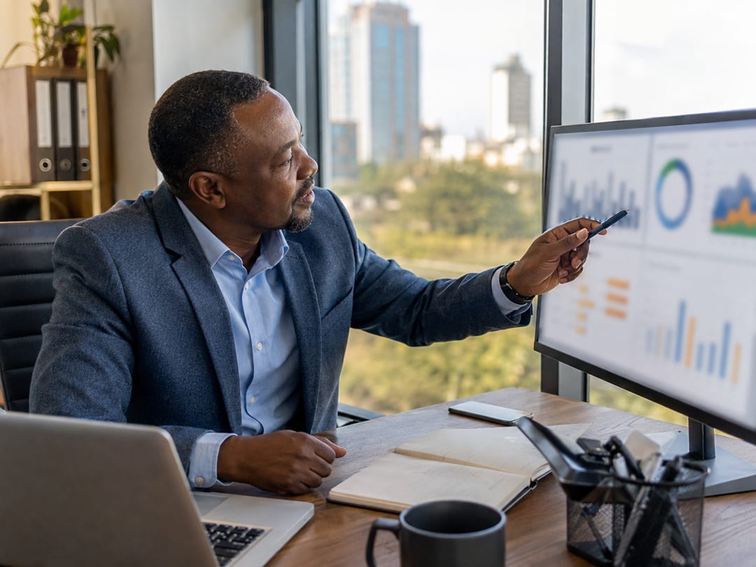 A fleet manager in a bright Nairobi office pointing a pen at charts on a large monitor, the city skyline through the window behind him.